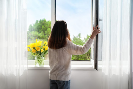 A woman looking out an open window in a home; there's a vase of yellow flowers on the window sill.